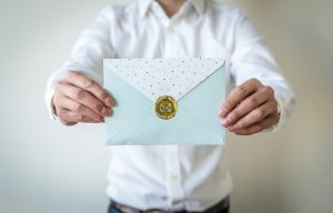 Man holding out a light blue envelope that has a gold circle sticker on the back that has "Happy Mail" embossed on it.